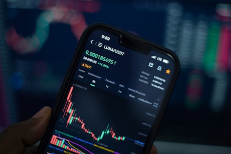 A close-up view of a smartphone displaying a cryptocurrency trading app with a graph showing the price movement of LUNA/USDT. The phone screen shows the current price, percentage change, and volume. The background is blurred with colorful digital charts, suggesting a trading environment.