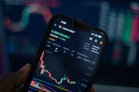 A close-up view of a smartphone displaying a cryptocurrency trading app with a graph showing the price movement of LUNA/USDT. The phone screen shows the current price, percentage change, and volume. The background is blurred with colorful digital charts, suggesting a trading environment.