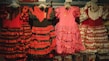 Traditional flamenco dresses hanging on display, featuring vibrant colors and ruffled layers. The dresses showcase polka dot patterns and are predominantly red, pink, and black with varying designs.