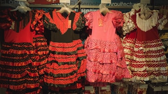 Traditional flamenco dresses hanging on display, featuring vibrant colors and ruffled layers. The dresses showcase polka dot patterns and are predominantly red, pink, and black with varying designs.
