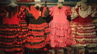 Traditional flamenco dresses hanging on display, featuring vibrant colors and ruffled layers. The dresses showcase polka dot patterns and are predominantly red, pink, and black with varying designs.