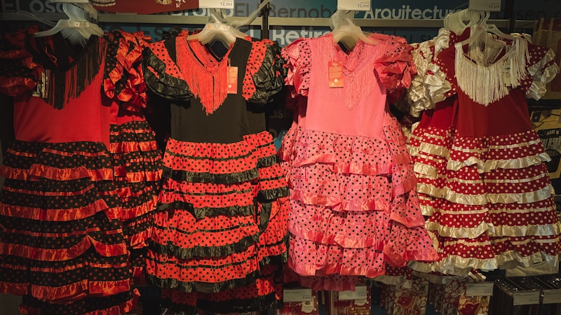 Traditional flamenco dresses hanging on display, featuring vibrant colors and ruffled layers. The dresses showcase polka dot patterns and are predominantly red, pink, and black with varying designs.