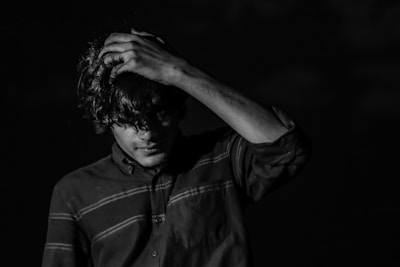 A person wearing a striped shirt holds their head with one hand, appearing contemplative or distressed. The setting is dark, with strong contrasts between light and shadow on the person's face and clothing.