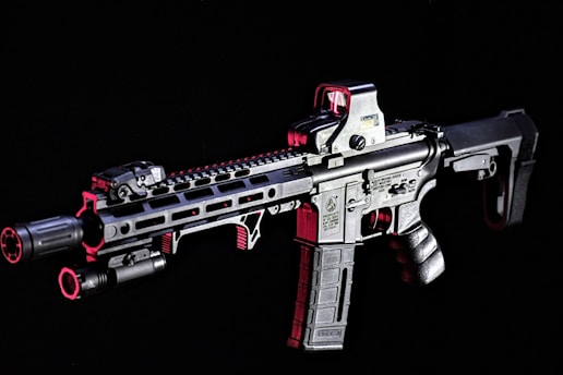 A modern rifle with a sleek design is set against a dark background. The gun features various attachments, including an optical sight on top and a flashlight mounted on the side. Metallic surfaces are highlighted with shades of red, adding contrast and depth to the appearance.