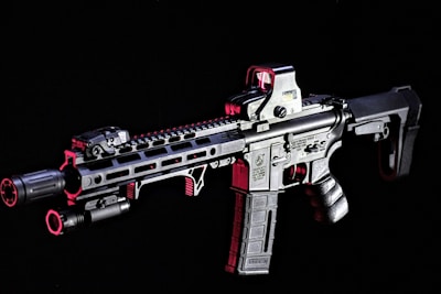 A modern rifle with a sleek design is set against a dark background. The gun features various attachments, including an optical sight on top and a flashlight mounted on the side. Metallic surfaces are highlighted with shades of red, adding contrast and depth to the appearance.