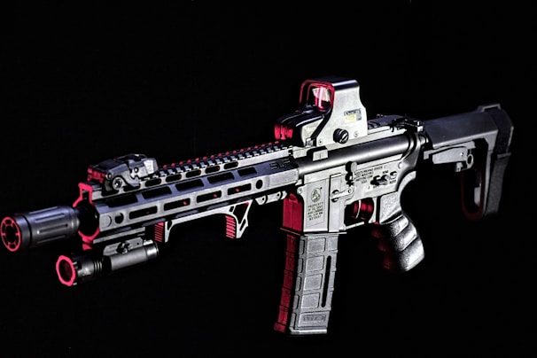 A modern rifle with a sleek design is set against a dark background. The gun features various attachments, including an optical sight on top and a flashlight mounted on the side. Metallic surfaces are highlighted with shades of red, adding contrast and depth to the appearance.