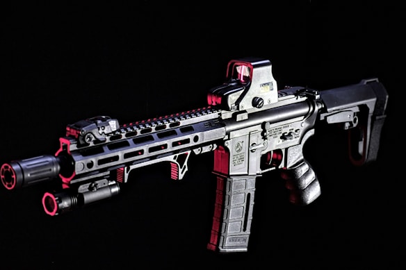 A modern rifle with a sleek design is set against a dark background. The gun features various attachments, including an optical sight on top and a flashlight mounted on the side. Metallic surfaces are highlighted with shades of red, adding contrast and depth to the appearance.