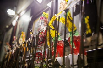 A collection of colorful traditional shadow puppets is displayed against a backdrop of framed art. The puppets feature intricate designs and vibrant colors, including reds, greens, and yellows, under bright lighting that accentuates their detail.