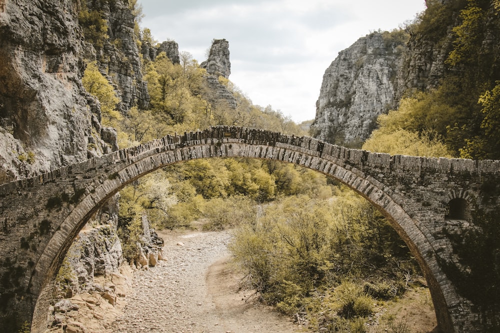 a stone bridge over a dirt road surrounded by mountains