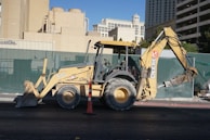 A yellow backhoe loader is parked on a street, with a large shovel on the front and an excavator arm on the back. The background includes a green construction fence and various buildings, including what appears to be hotel architecture.