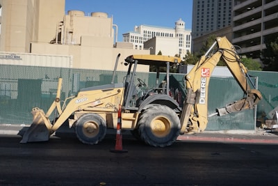 A yellow backhoe loader is parked on a street, with a large shovel on the front and an excavator arm on the back. The background includes a green construction fence and various buildings, including what appears to be hotel architecture.