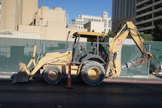 A yellow backhoe loader is parked on a street, with a large shovel on the front and an excavator arm on the back. The background includes a green construction fence and various buildings, including what appears to be hotel architecture.
