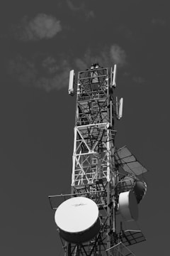 A large metal communication tower equipped with multiple satellite dishes and antennas, set against a cloudy sky.