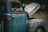 A weathered, old pickup truck with its rusted blue and gray body shows signs of age and use. The hood is open, revealing a glimpse of its engine. A small American flag is visible near the window, adding a touch of color and patriotism.