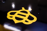 A stylized yellow bee logo is prominently featured against a glossy black background. The bee design is minimalist, using simple geometric shapes to convey the insect's form. The surface reflects light, adding a shiny texture to the overall appearance.