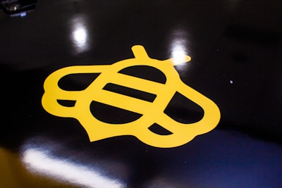 A stylized yellow bee logo is prominently featured against a glossy black background. The bee design is minimalist, using simple geometric shapes to convey the insect's form. The surface reflects light, adding a shiny texture to the overall appearance.
