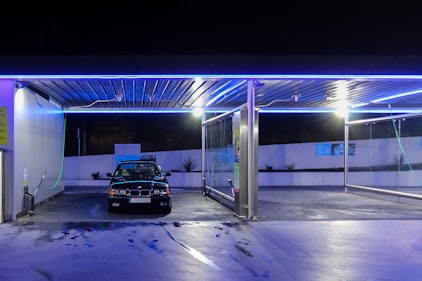 A car is parked in an automated car wash station at night. The station is illuminated with blue neon lights, creating a modern, sleek atmosphere. The ground appears wet, indicating recent washing activity. The surrounding area is clean and well-maintained, with a white wall in the background adorned with some plants.