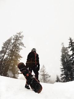 A person dressed in winter gear stands on a snowy slope with a snowboard, surrounded by tall evergreen trees. The sky is overcast, and snowflakes are visibly falling, creating a wintry atmosphere.