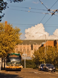 A city street scene features a public transport bus moving towards the viewer, surrounded by leafy trees. In the background, a large, historic building with intricate architecture and a statue on its roof dominates the scene, under a blue sky with fluffy clouds. Cars are parked along the street, and overhead cables stretch across the scene.