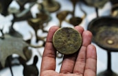 A hand holding a vintage coin with engravings, surrounded by blurred bronze artifacts in the background.