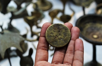 A hand holding a vintage coin with engravings, surrounded by blurred bronze artifacts in the background.