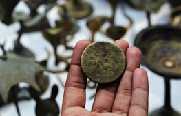 A hand holding a vintage coin with engravings, surrounded by blurred bronze artifacts in the background.