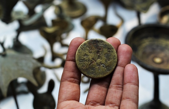 A hand holding a vintage coin with engravings, surrounded by blurred bronze artifacts in the background.