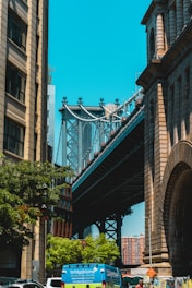 A bridge seen between two tall buildings, with a clear blue sky in the background. The structures are surrounded by trees and there is a blue van on the street below, with additional vehicles partially visible.