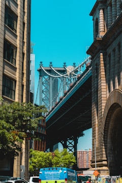 A bridge seen between two tall buildings, with a clear blue sky in the background. The structures are surrounded by trees and there is a blue van on the street below, with additional vehicles partially visible.