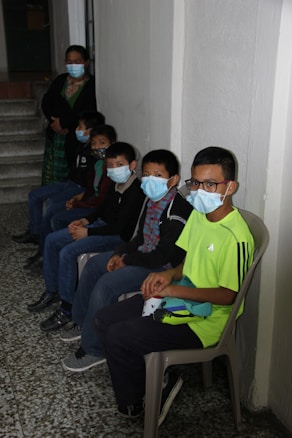 Five children wearing face masks are seated in a line against a wall indoors, with one adult standing beside them. They are sitting on plastic chairs on a terrazzo floor.