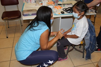 A woman and a young girl are wearing face masks in a medical setting. The woman is kneeling and interacting with the girl, who is sitting on a stool. Medical supplies and equipment are visible on a table in the background.
