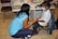 A woman and a young girl are wearing face masks in a medical setting. The woman is kneeling and interacting with the girl, who is sitting on a stool. Medical supplies and equipment are visible on a table in the background.