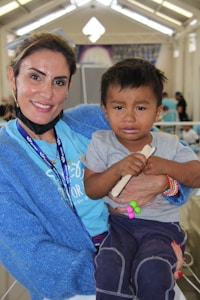 A woman with short brown hair smiles warmly while holding a young child who appears to be crying. The woman is wearing a blue knitted sweater and a blue shirt, with a lanyard around her neck. The child is wearing a gray shirt and dark pants, holding a small colorful toy and a stick. The background shows an indoor setting with a high ceiling and some other people visible in the distance.