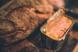 A rustic loaf of bread with a crusty texture sits next to an open can of processed meat, likely pate or luncheon meat. The scene is warmly lit, giving it a cozy and vintage feel.