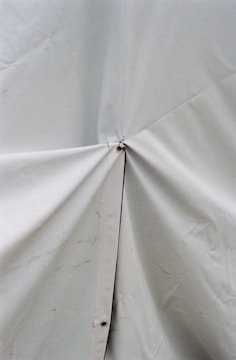 A close-up view of a light-colored tarp or fabric material with a prominent central joining point secured by a metal or plastic fastener. The surface shows some wrinkles and slight discoloration.