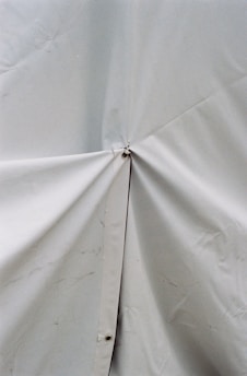 A close-up view of a light-colored tarp or fabric material with a prominent central joining point secured by a metal or plastic fastener. The surface shows some wrinkles and slight discoloration.