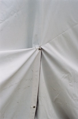 A close-up view of a light-colored tarp or fabric material with a prominent central joining point secured by a metal or plastic fastener. The surface shows some wrinkles and slight discoloration.