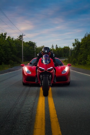A motorcycle aligned directly in front of a red sports car on an empty road, with both vehicles facing forward. The motorcyclist is wearing a black helmet and matching gear. The scene is dynamic, with a background of green trees lining the road and a picturesque sky above.