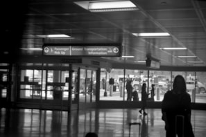 A black and white photo of an airport terminal with a sign directing to Passenger Pick Up, Ground Transportation, and Information. Silhouettes of people with luggage are visible inside the terminal. Outside, through large glass windows, parked vehicles and more people can be seen.
