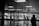 A black and white photo of an airport terminal with a sign directing to Passenger Pick Up, Ground Transportation, and Information. Silhouettes of people with luggage are visible inside the terminal. Outside, through large glass windows, parked vehicles and more people can be seen.