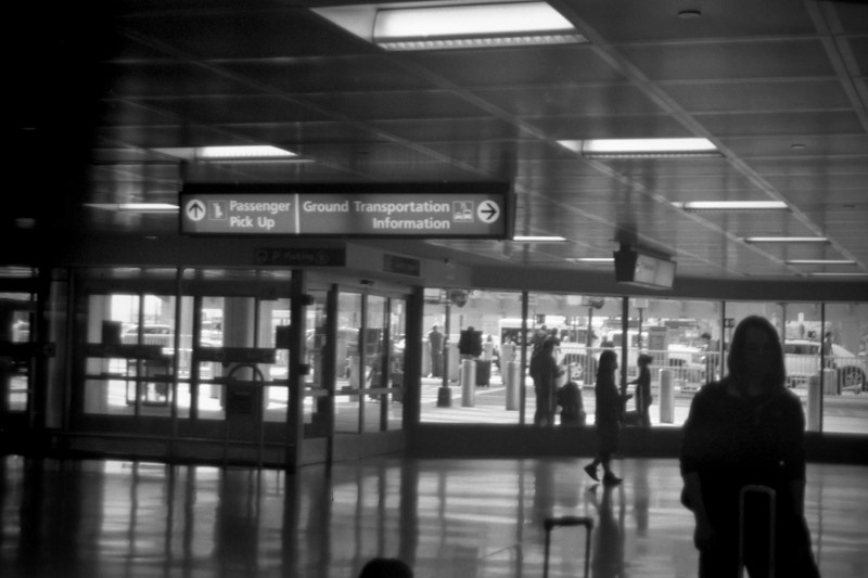A black and white photo of an airport terminal with a sign directing to Passenger Pick Up, Ground Transportation, and Information. Silhouettes of people with luggage are visible inside the terminal. Outside, through large glass windows, parked vehicles and more people can be seen.