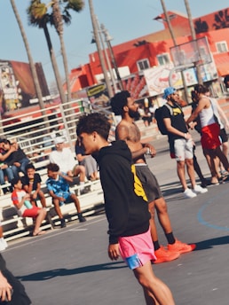 A group of people gathered in an outdoor area, possibly a basketball court near a beach. There are palm trees in the background and colorful buildings with murals. The scene includes several young men, some wearing athletic attire. A few individuals are seated on bleachers.