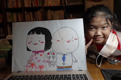 A smiling child with a face mask partially off leans next to a drawing of two cartoon characters holding hands. The characters are dressed in colorful clothing, one in a floral dress and the other in a shirt and tie. Bookshelves are visible in the background, suggesting a cozy interior setting.