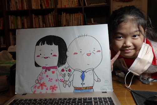 A smiling child with a face mask partially off leans next to a drawing of two cartoon characters holding hands. The characters are dressed in colorful clothing, one in a floral dress and the other in a shirt and tie. Bookshelves are visible in the background, suggesting a cozy interior setting.