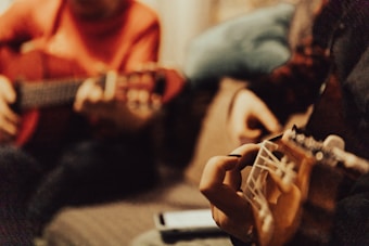 Two people are sitting closely together, each playing a guitar. The focus is on their hands and guitars, with a cozy and intimate atmosphere suggested by the warm colors and blurred background.