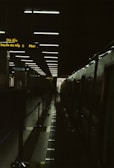 A dimly lit train station platform with overhead lights reflecting on a glossy floor. The platform is flanked by a stationary train on the right side, and a series of illuminated signs with yellow text. A doorway at the end of the platform lets in a small amount of natural light.