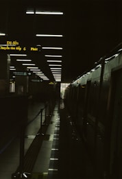 A dimly lit train station platform with overhead lights reflecting on a glossy floor. The platform is flanked by a stationary train on the right side, and a series of illuminated signs with yellow text. A doorway at the end of the platform lets in a small amount of natural light.