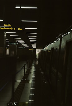 A dimly lit train station platform with overhead lights reflecting on a glossy floor. The platform is flanked by a stationary train on the right side, and a series of illuminated signs with yellow text. A doorway at the end of the platform lets in a small amount of natural light.