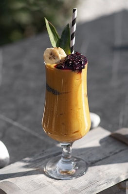 A vibrant, tall smoothie in a clear glass, topped with banana slices, pineapple leaves, and a dollop of dark-colored puree or jam, with a black and white striped straw inserted in it. The smoothie itself has a rich, yellow-orange hue and appears creamy. The background suggests a wooden table surface with soft, natural lighting enhancing the colors and textures.