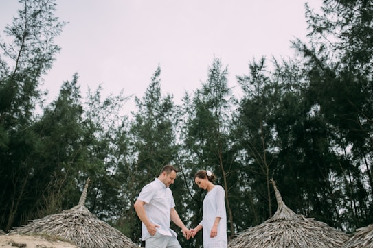 A couple is standing outdoors surrounded by tall trees and traditional thatched-roof structures. They are holding hands and appear to be looking down. The scene is serene and intimate, set against a backdrop of dense green foliage.